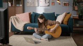Woman is playing guitar, sitting on the floor. Young musician is using online video lesson to improve skills. Guitarist is practicing melody. Cozy living room, remote learning, creative hobbies.