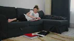 Young woman makes notes in notebook from computer, is lying on the bed at home and laptop is on the floor. Working on computer. Concept of remote work, telework. Concentrated she writes in notebook.
