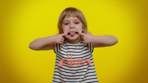 Portrait of cheerful funny blonde child girl 5-6 years old showing tongue making faces at camera, fooling around, joking, aping with silly face, teasing. Yellow studio background. Teen kid children