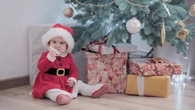 authentic cute happy Joy chubby infant girl wearing santa hat and red dress smile play have fun celebrating new year festive atmosphere near christmas tree at home. Childhood, hollyday, winter concept