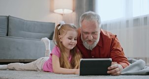 Happy family adult parent dad and small daughter having fun using digital tablet lying on floor at home. Cute child girl learning technology talking with father teaching kid look at pad screen at home