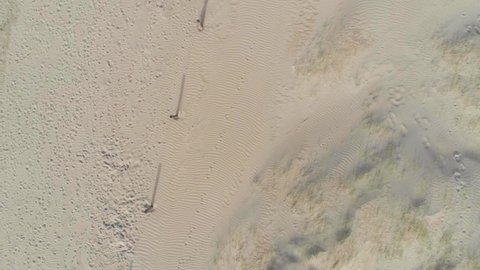 Long fence post shadows stretch out across a smooth sandy beach covered in footprints, aerial god view