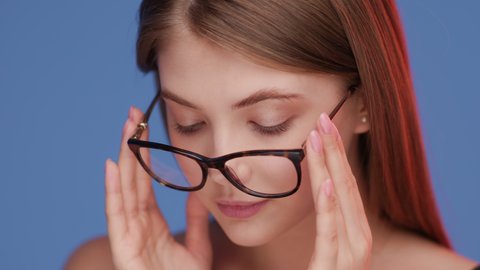 Young woman puts on glasses and looks at camera. Positive portrait of sexy female model with nude make-up close-up. Eyes optics for good vision and normal sight. Advertising of eyewear shop indoors