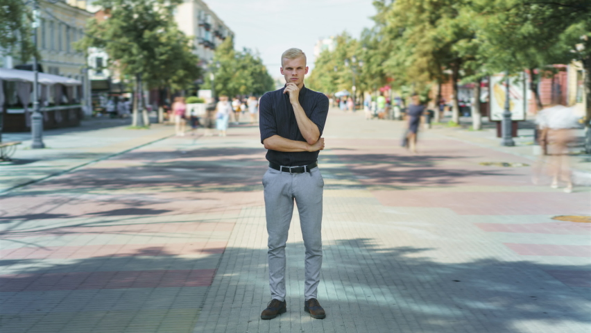 Zoom-in time lapse of pensive guy in elegant clothing standing outside pedestrian street looking at camera while men and women are passing by