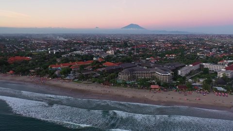 Dramatic aerial drone footage of the sunset over the famous Kuta beach in Seminyak lined with beach bars and Mt Agung volcano in the background in Bali, Indonesia. 