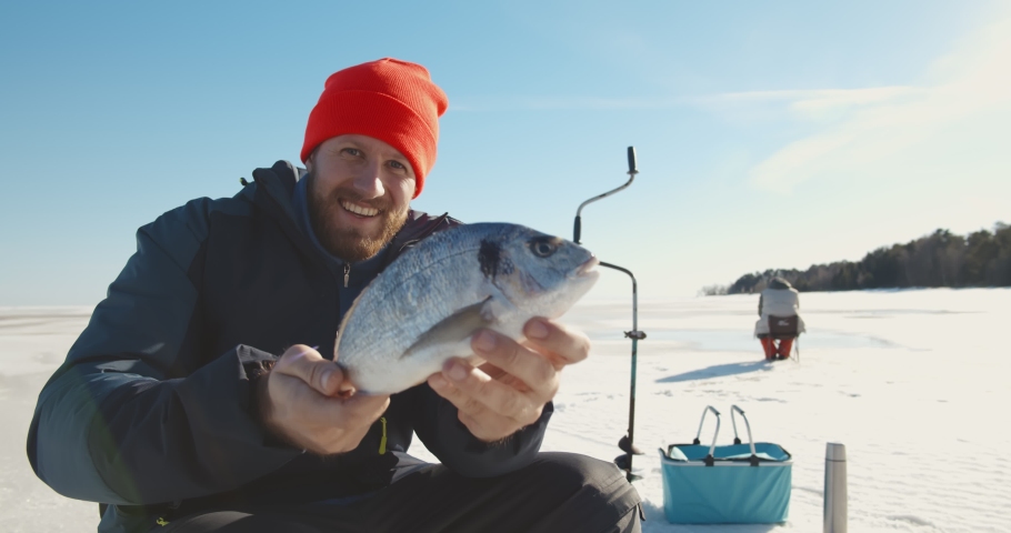 Happy bearded fisherman holding fish and smiling at camera enjoying winter fishing. Portrait of young fisher catching fish in hole on frozen lake or river in winter