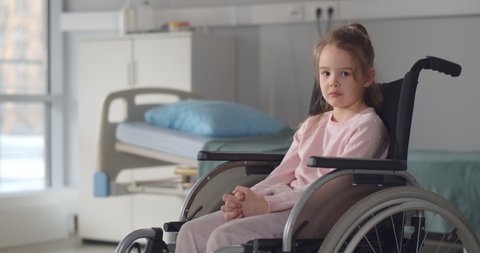 Sad little girl in a wheelchair in hospital ward looking at camera. Portrait of upset and lonely disables child patient sitting in wheelchair at pediatrics department