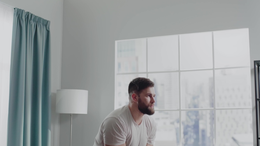 Muscular guy exercises dumbbell snatch with cast iron weight in hands standing at home back to window in morning slow motion