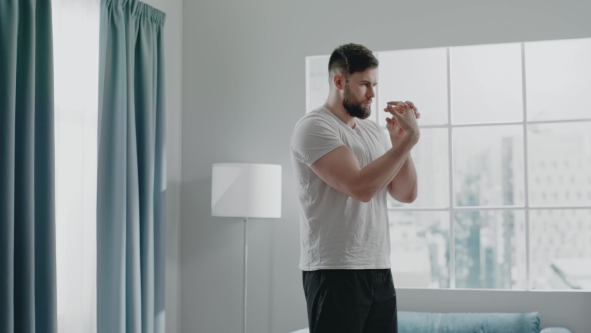 Experienced bearded martial artist in t-shirt and shorts practices wrists warmup standing against large window in gym slow motion