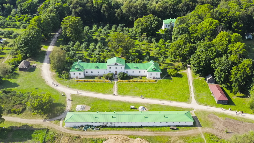 Village in the Shchyokinsky district of the Tula region - Yasnaya Polyana, Russia. The Tolstoy House Museum, a white-painted manor house with a distinctive green metal roof, sits at the center of the estate surrounded by manicured lawns and dense woodland. The house is one of two surviving wings of the original Volkonsky family manor built in the early 19th century, where Leo Tolstoy was born in 1828 and where he wrote both War and Peace and Anna Karenina. A long rectangular farm building with a matching green roof stands in the foreground, while winding dirt paths and scattered outbuildings extend across the broad, gently sloping grounds. The estate is set amid extensive forests and rolling hills under a bright summer sky, captured from a high aerial perspective that reveals the full layout of the historic property. The scene conveys a sense of pastoral tranquility and cultural heritage, with the lush green landscape dominating the composition and the architectural ensemble providing a focal point of literary significance.