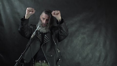Bearded hairy rock musician in leather jacket blows a kiss, holding fists up and leaves the stage on concert with black curtain background