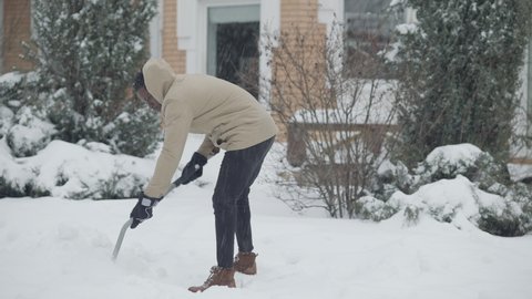 Wide shot of young African American man cleaning snow under snowfall outdoors. Confident concentrated handsome guy working on snowy day with shovel. Lifestyle and cold concept