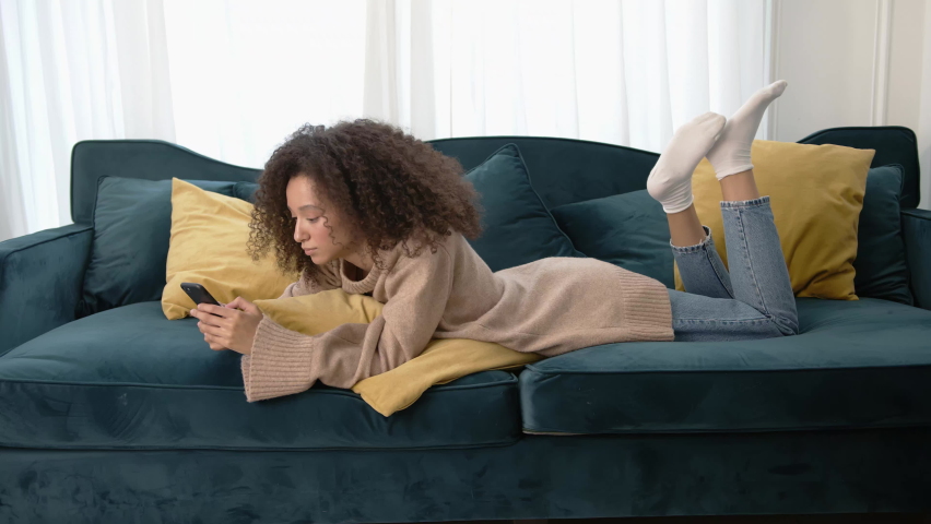 African woman using phone, chilling on a long sofa in home living room. Young curly hair woman in beige sweater, smiling and laughing, typing on the mobile phone