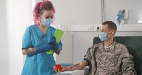 Nurse in protective mask talking to military servant sitting on chair and receiving blood transfusion at hospital. Medical worker with clipboard talking to soldier blood donor