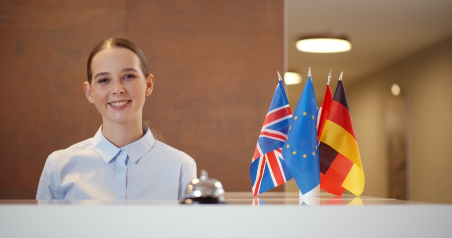Portrait of beautiful young woman hotel receptionist. International hotel worker smiling at camera standing at front desk with german. England and us flags