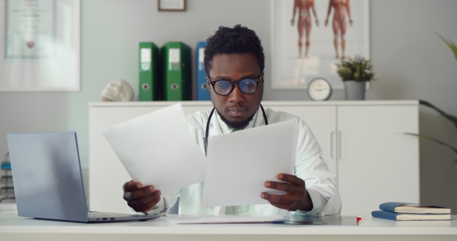 African man doctor studying medical report of patient sitting at desk in office room. Young afro-american practitioner doing paperwork in hospital office | Shutterstock HD Video #1071214249