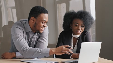 Friendly African American expert educate intern, helps millennial lady girl. Young man, woman work together at computer on financial statistics analyzing charts project data collaborate successfully