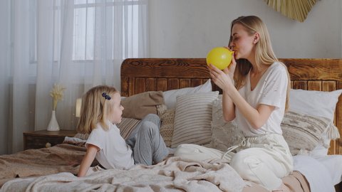 Little girl child daughter baby kid sitting with loving mother babysitter on bed in cozy bedroom play looks how mom inflates blowing air in yellow balloon family laughs enjoying holiday time together