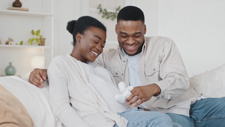 African American couple family future expecting parents sitting on couch afro black husband man stroking wife pregnant belly with cute small childish child white shoes hugging embracing beloved woman Royalty-Free Stock Footage #1071217363
