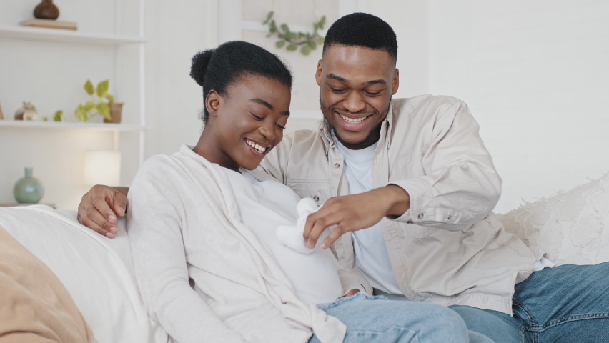 African American couple family future expecting parents sitting on couch afro black husband man stroking wife pregnant belly with cute small childish child white shoes hugging embracing beloved woman | Shutterstock HD Video #1071217363