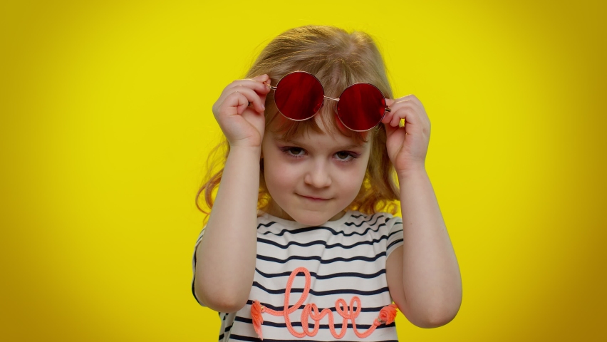 Playful happy little blonde teen kid child girl in sunglasses blinking eye, looking at camera with toothy smile, winking and flirting, expressing optimism. Young children on yellow studio background | Shutterstock HD Video #1071218356