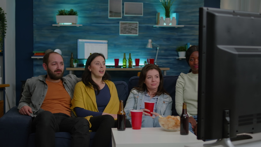 Multiracial friends laughing smiling having fun together while sitting on couch in front of TV watching comedy movie. Group of mixed race people enjoying free time drinking beer eating snacks Royalty-Free Stock Footage #1071223096