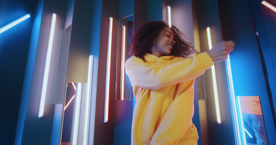 Active urban african american black woman in yellow hoodie dancing by neon lights background in disco club. Cool talented mixed race female dancer feeling free moves rhythmically to hip-hop music beat | Shutterstock HD Video #1071226297