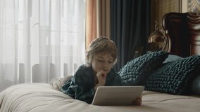 Child smiling chatting online by tablet lying on bed. Using handheld device. Communication via internet distantly. Boy having a video call at home.