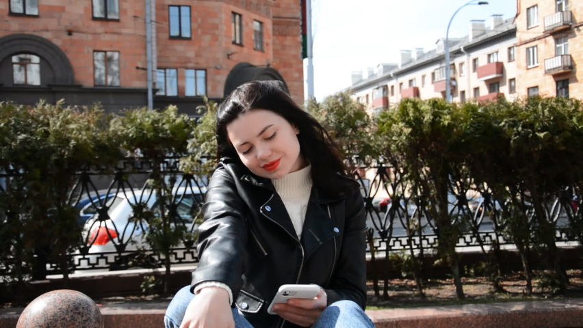 Young caucasian pretty woman sitting on bench at bus stop using smartphone with internet for texting online message
