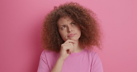 Beautiful thoughtful woman with curly hair rubs chin thinks deeply chooses or ponders plan searches for solution feels hesitant dressed in casual clothes isolated over pink background has pensive face