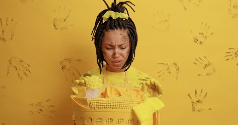 Displeased Afro American woman smirks face looks with aversion at dirt wears rubber gloves poses near basket full of cleaning supplies has dreadlocks hairstyle feels unhappy and tired of housework