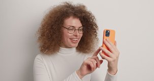 Overjoyed glad European woman with curly hair waves hello in smartphone greets someone feels very happy wears casual turtleneck isolated over white background enjoys friendly videoconference