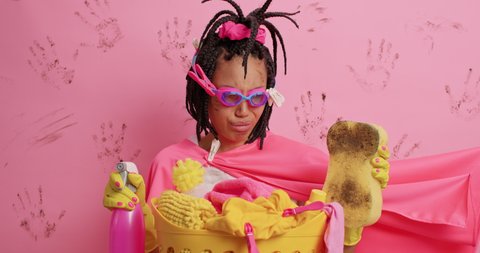 Spring cleaning concept. Displeased female domestic helper busy doing clean up smirks face with aversion holds dirty sponge and detergent dressed like superhero isolated over pink background