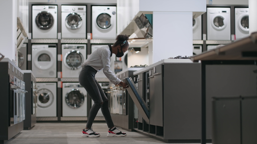 Young black woman with face mask is viewing dishwasher in shop, mall with department of home appliances, choosing equipment for kitchen | Shutterstock HD Video #1071240850
