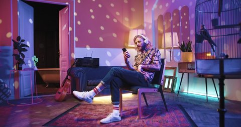 Young stylish Caucasian cool man sit on chair lin vintage apartment listening to music in headphones at home in room with warm light and typing on cellphone searching internet surfing social network