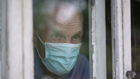 Old male person wearing medical face mask looking through window.