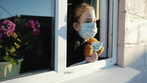 Quarantine, threat of coronavirus. Sad child and his teddy bear. Bored girl wears medical mask in home quarantine coronavirus looks out of the window. Epidemic covid 19 prevention concept.