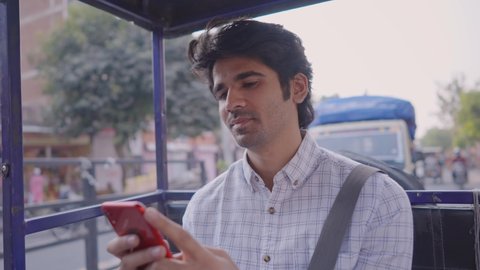 close shot of a young attractive Indian male office employee is smiling and using a mobile phone to type a text message in a moving an auto-rickshaw through a busy city market road  
