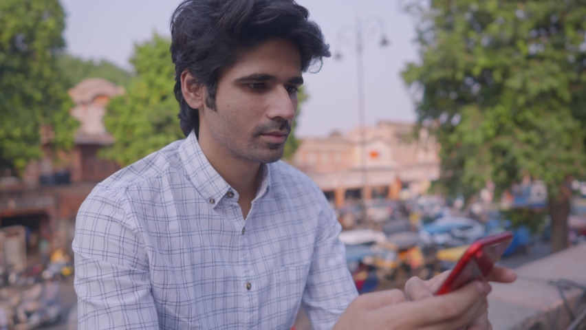 Close shot of an attractive young Indian male office worker sitting on a rooftop of a building smiling looking at the mobile phone with busy city market road and traffic in the background. Royalty-Free Stock Footage #1071261733