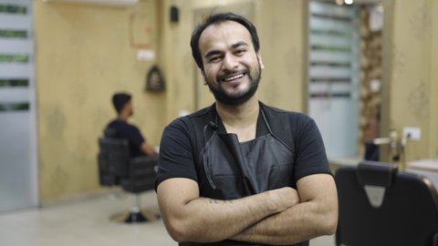 close up shot of a young male Indian Asian barbershop owner standing and smiling with crossed arms in a salon wearing a spa apron and looking at the camera. 