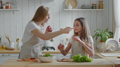 Mother and daughter cook natural vegetable salad, mommy feeding baby from wooden spoon, girl eating delicious dish rejoices gives high five to woman jumping with happiness, family hugging in kitchen