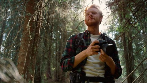 Smiling photographer looking at photos on professional camera outdoor. Redhead man with photo camera shooting in fairytale forest. Handsome guy looking around natural landscape