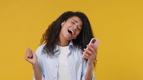 Fun happy young african woman in blue white t shirt listen music in earphones dance sing song hold mobile cell phone fooling around have fun enjoy relax isolated on yellow color wall background studio