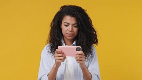 Gambling young african woman curly hair 20s years old wears blue white t shirt using play racing on mobile cell phone hold gadget smartphone for pc video games isolated on yellow color wall background
