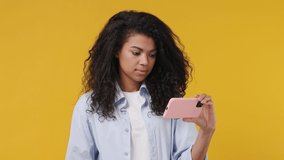 Young african woman wears blue white t shirt hold use mobile cell phone swipe browsing doing online shopping order order delivery to home booking tour isolated on yellow color wall background studio