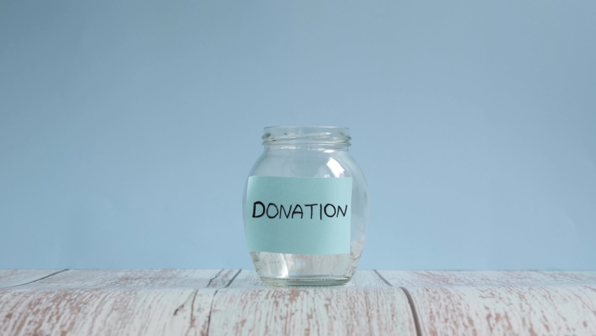 Hand throwing coins in transparent jar with inscription donation. Coins are falling down inside glass jar. Saving money for donation | Shutterstock HD Video #1071278110