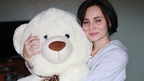 Portrait of happy woman with short hair hugging a big teddy bear at home. Mother touching plush toy and closing eyes. Sleeping concept