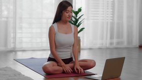 A beautiful Asian woman sitting on a yoga mat looking at a laptop screen with a teacher teaching yoga online. She does meditation and lotus pose as the starting point of yoga.