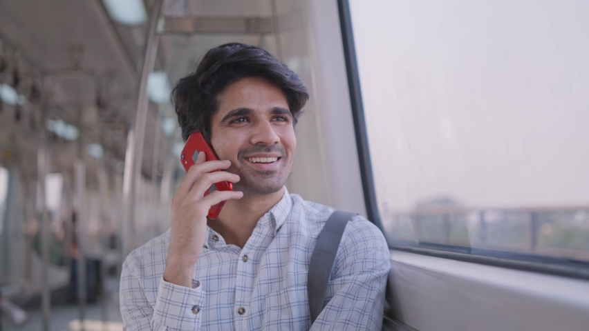 A young attractive Indian cheerful male corporate office employee in formal wear sitting and speaking on a mobile phone on a moving modern city metro train while going to the workplace Royalty-Free Stock Footage #1071290236