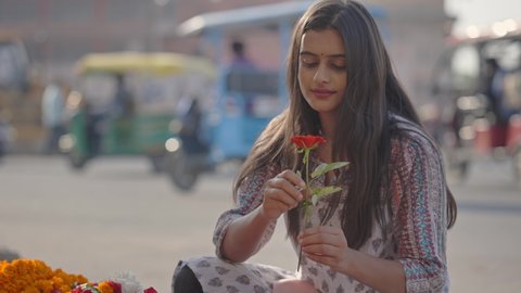A young attractive Indian Asian female in a traditional salwar kameez dress is smelling and buying a red rose from a colorful flower shop or a street vendor next to a busy road 